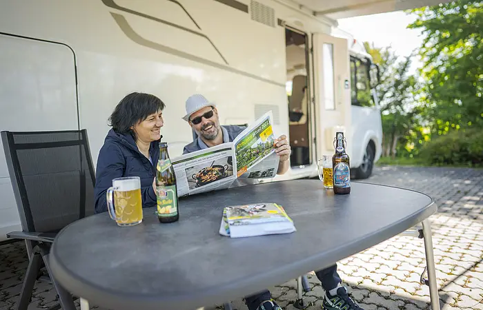 Pärchen auf dem Reisemobilstellplatz sitzt vor dem Wohnmobil und liest die NPA- Zeitung. Sind im Fokus des Bildes. Beide lachen über den Inhalt .