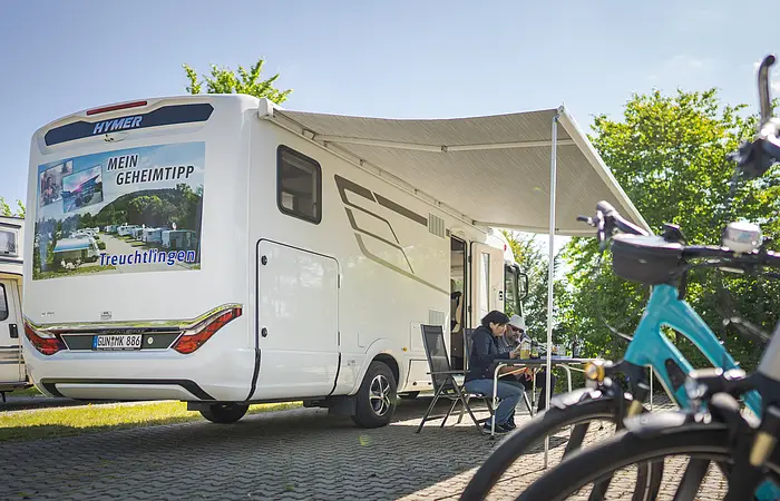 Wohnmobil steht mit ausgefahrener Markise am Stellplatz. Im Fokus die Räder zweier abgestellter Fahrräder. Wohnmobil hat große Werbung für den Stellplatz hinten drauf.