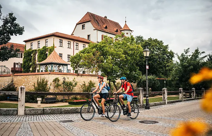 Zwei Personen mit Fahrradhelmen fahren auf einem gepflasterten Weg vor einem historischen Gebäude mit Bäumen.