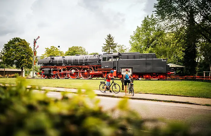 Dampflokomotive auf Gleis mit zwei Radfahrern davor, umgeben von Bäumen und Wiese bei bewölktem Himmel.