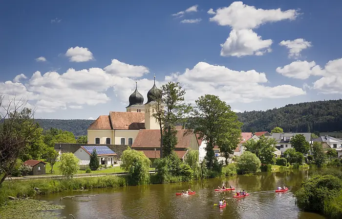 Auf der Altmühl paddeln fünf rote Kanus an Kottingwörth mit ihrer Kirche und deren zwei Kirchtürmen vorbei.