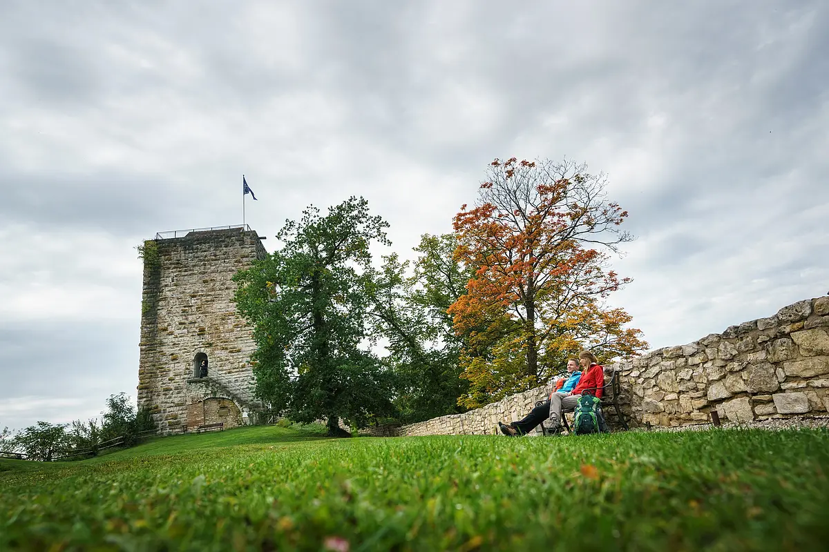 Wanderer an der Burg Pappenheim Zwei Personen sitzen auf einer Bank neben einer Steinmauer vor einem Turm und herbstlich gefärbten Bäumen.