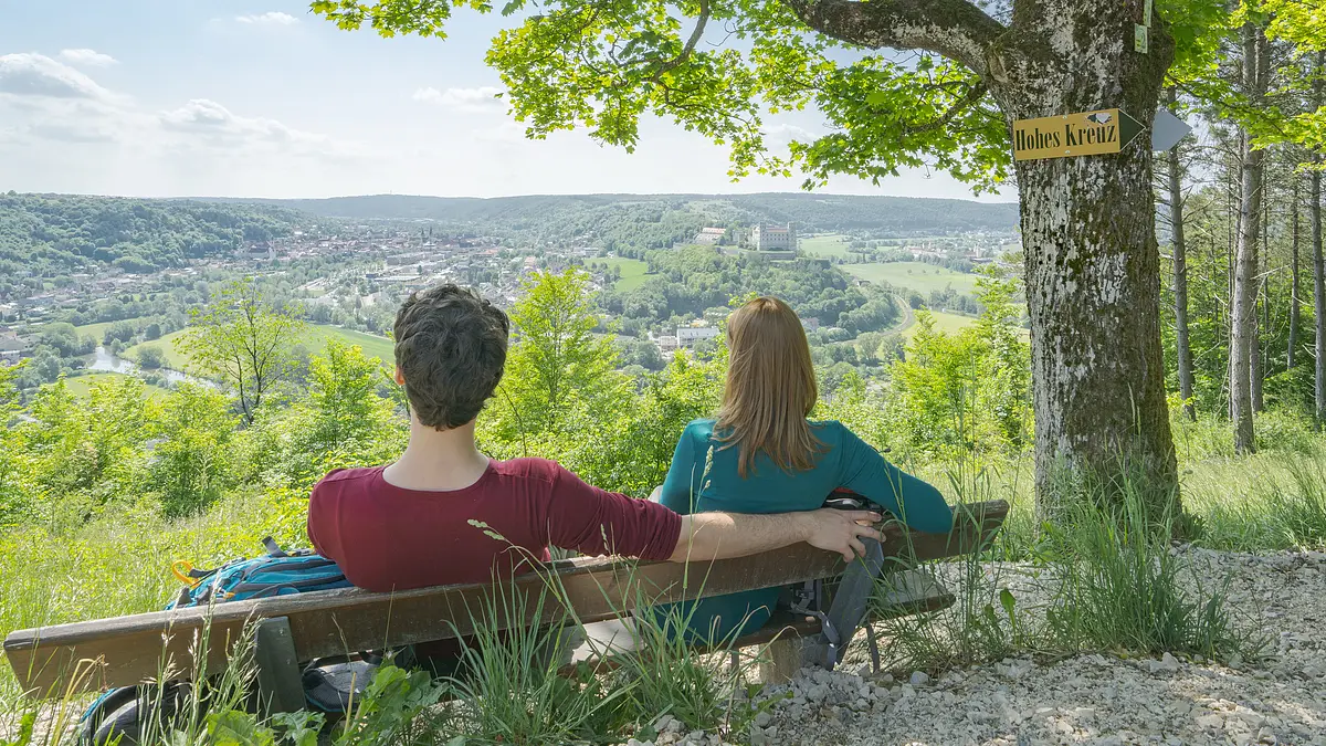 Altmühltal-Panoramaweg (Eichstätt) Zwei Personen sitzen auf einer Bank unter Baum mit Schild „Hohes Kreuz“ und blicken auf ein Tal mit Stadt und Hügeln.