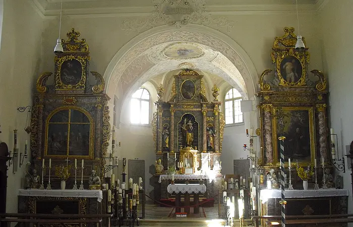 Innenraum einer Kirche mit barocken Altären, verziert mit Gemälden und goldenen Ornamenten. Sonnenlicht fällt durch ein Fenster.