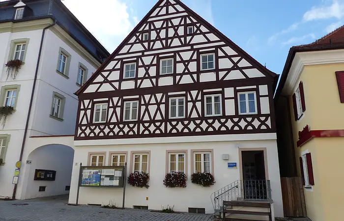 Tourist-Information der Stadt Monheim/Monheimer Alb: ein Fachwerkhaus mit drei Stockwerken, weißen Wänden und braunen Balken. Davor steht eine Sitzbank und ein Schaukasten auf einem gepflasterten Platz. Links davon ein weißes dreistöckiges Gebäude. Rechts davon ein gelbes zweistöckiges Gebäude mit roten Fensterläden.