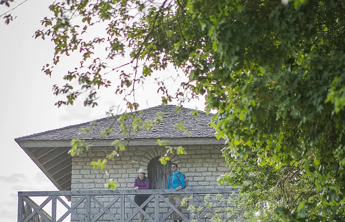 Eine Frau mit Strohhut und ein Mann mit blauer Jacke stehen an dem hölzernen Geländer oben auf dem Limesturm. Hinter ihnen ist eine hölzerne Tür mit der man ins innere des Turms gelangt. Der Turm hat weiter unten noch ein kleines Fenster und links sind einige Steinstufen. Rechts wird der Turm von grünen Blättern der Bäume eingerahmt.