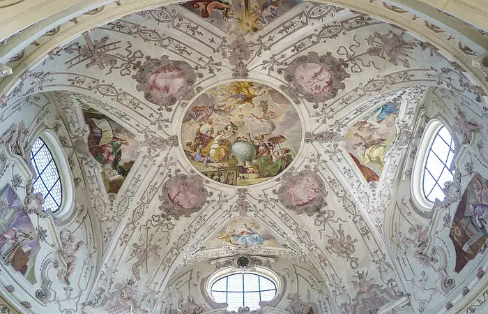 Deckenfresko mit religiösen Szenen und Ornamenten in einer Kirche, umgeben von Fenstern und Stuckverzierungen.
