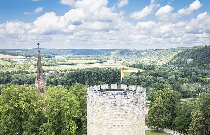 Runder Steinturm mit Fahne auf einer Wiese, im Hintergrund eine Kirche und bewaldete Hügel.