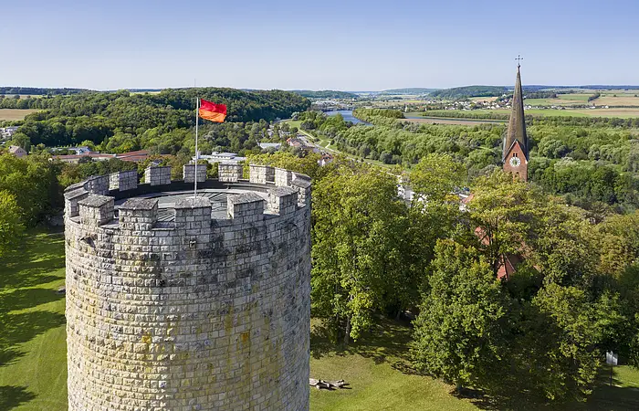 Runder Steinturm mit deutscher Flagge neben Kirchturm in grüner Landschaft mit Fluss und Feldern im Hintergrund.