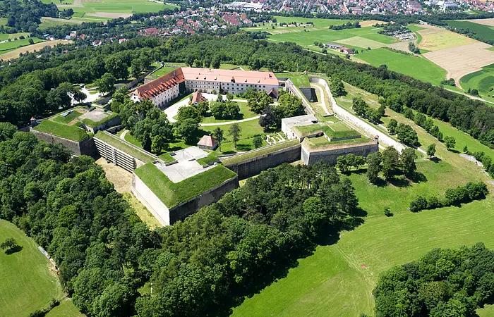 Luftaufnahme einer Festungsanlage. Eine Burg umgeben von einer Mauer in der Form eines fünfzackigen Sterns. Um die Festungsanlage Wälder und Felder.