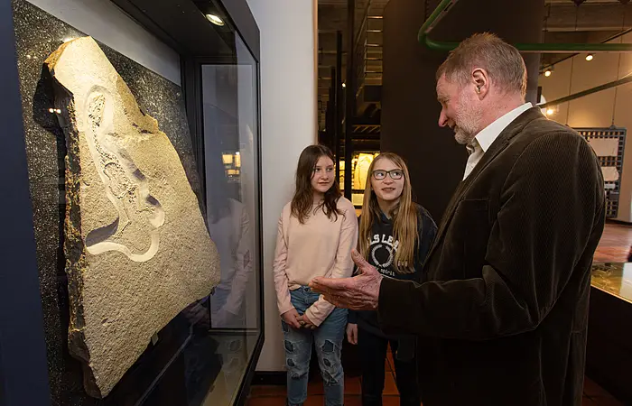 Zwei Mädchen, eine blond und die andere braunhaarig stehen neben einer Versteinerung und schauen interessiert zu dem Mann mit braunem Jackett, der ihnen mehr zu den Fossilien erzählt.