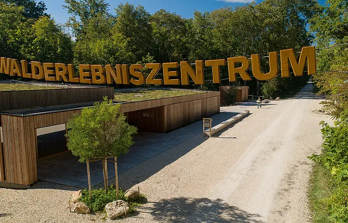 Am Walderlebniszentrum wird man am Eingang mit dem großen Schriftzug aus Holz empfangen. Darunter sieht man eine Holzhütte und rechts geht ein Feldweg in den Wald.