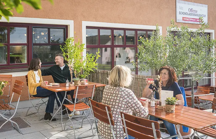 Außenbereich eines Cafés mit zwei Tischen, an denen vier Personen Kaffee trinken und sich unterhalten.