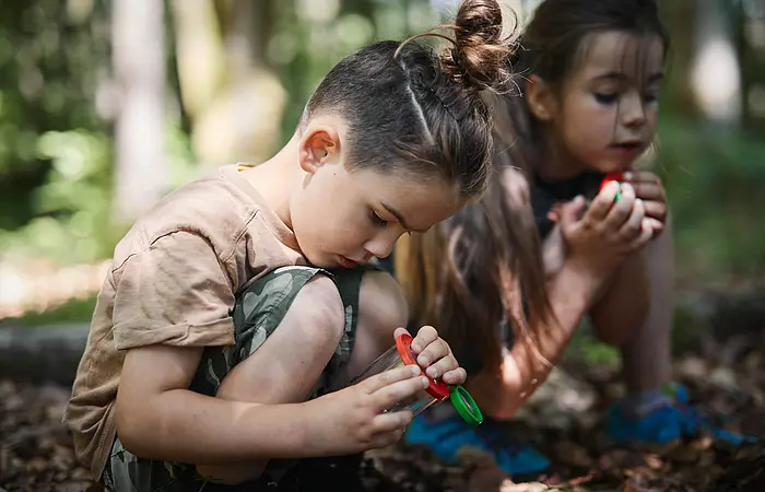 Zwei Kinder sitzen am Waldboden und halten sehr interessiert etwas fest, dass sie zudem gebahnt anschauen.