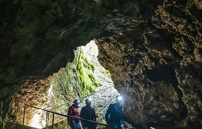 Drei Wanderer steigen die Treppenstufen zur Höhle hinab. Der Vorderste ist mit einer Stirnlampe ausgestattet und leuchtet ihnen den Weg aus. Licht kommt nur vom Eingang in die Höhle.
