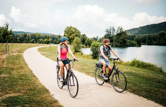 Eine Frau mit roten Shirt und blauem Fahrradhelm radelt neben einem Mann mit grauem Shirt und roten Helm. Rechts von ihnne liegt der Kratzmühlsee. Der Radweg ist von grünen Wiesen gesäumt. Der Himmel ist an diesem Tag etwas bewölkt.