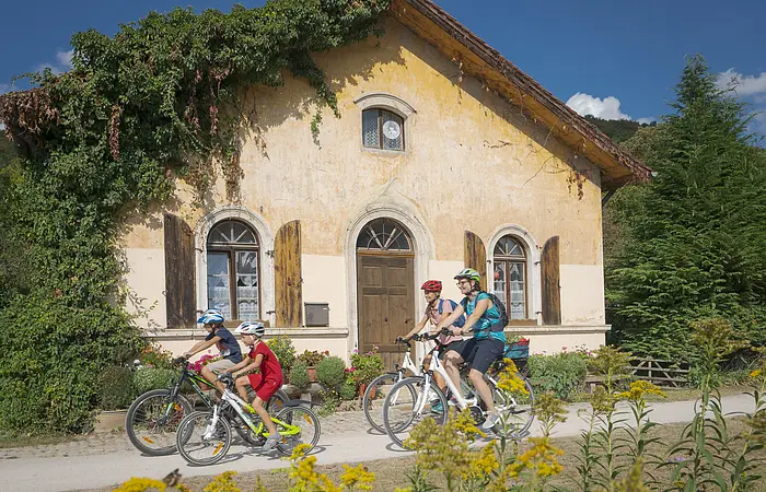 Eine Familie radelt auf dem Altmühltal-Radweg am Schleusenhäuschen vorbei. Die Radler haben alle einen Fahrradhelm auf. Das Häuschen ist auf der linken Seite etwas vom Effeu eingewachsen. Die Fenster haben braune Fensterläden.