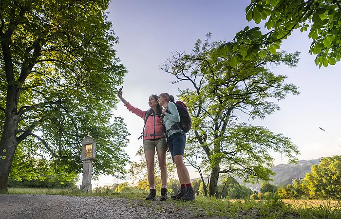 Zwei Frauen in rosafarbenen und hellblauem Shirt, sowie kurzen Hosen stehen eng beieinander am Frauenberg und die Frau in rosa, hält ihr Handy für ein gemeinsames Selfie in die Höhe. Ein großer Baum, sowie ein Marterl sind vor den Zweien zu erkennen. Im Hintergrund der zwei Frauen sind vereinzelt Bäume zu sehen , aber auch ein wolkenloser Himmel.