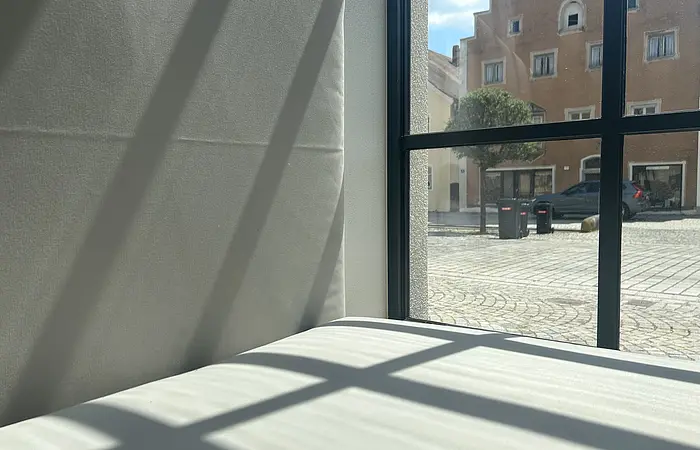 Fenster mit schwarzen Rahmen, durch das Sonnenlicht Schatten auf eine weiße Wand und Fläche wirft, draußen Gebäude und Pflastersteinweg sichtbar.