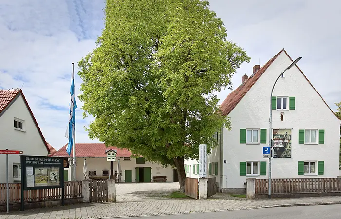Der Eingangsbereich des Museums mit seinen Gebäudlichkeiten. Links ist zudem eine Infotafel und eine Fahne angebracht. Der Blick in den Hof zeigt ein weiteres Gebäude und einen großen Baum.