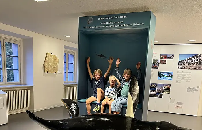 Drei Kinder sitzen vor einer blauen Museumsvitrine mit Text, davor ein großes Modell eines urzeitlichen Meeresreptils.