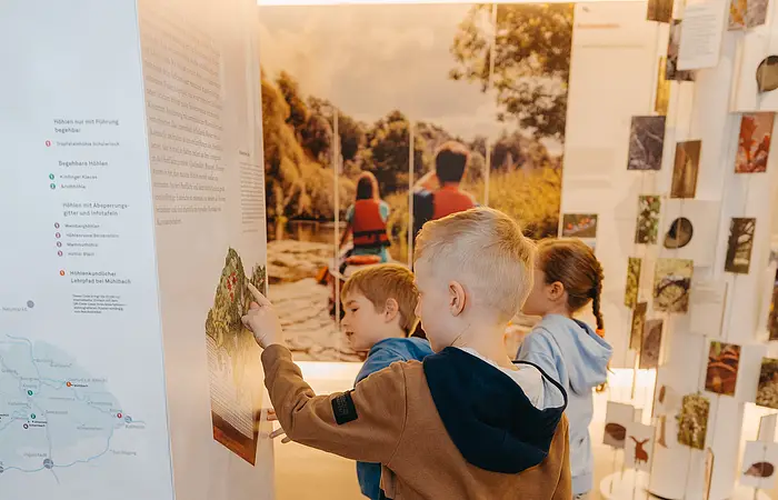 Drei Kinder betrachten eine Ausstellungstafel in einem Museum mit Natur- und Landschaftsbildern.