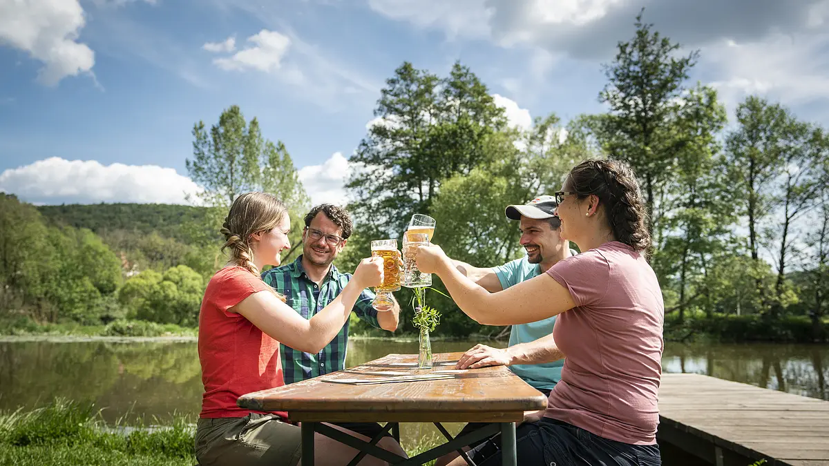 Vier Personen stoßen mit Biergläsern an einem Holztisch im Freien an, umgeben von Bäumen und einem See.