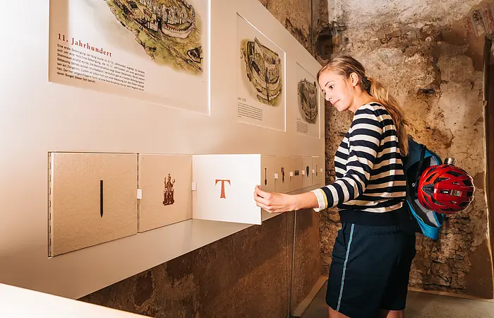 Frau in gestreiftem Pullover öffnet Klappe an Ausstellungstafel mit historischen Texten und Bildern.