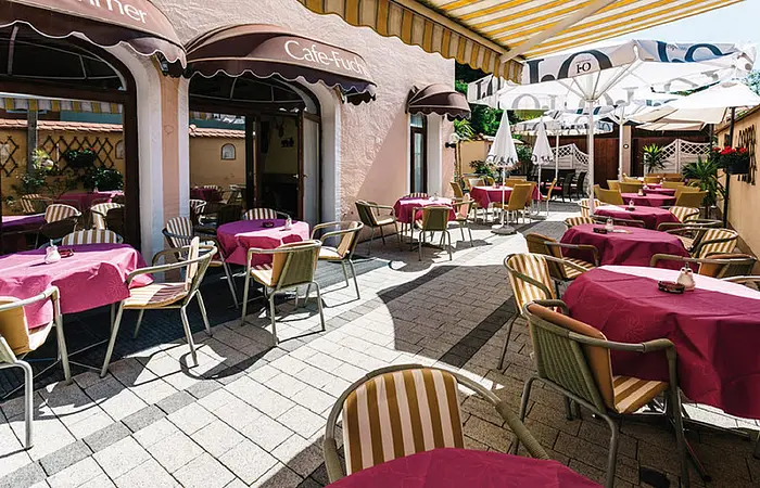 Das Bild zeigt den Außenbereich des "Café Fuchs". Die Terrasse ist mit Tischen und Stühlen ausgestattet, die mit pinken Tischdecken und gestreiften Sitzpolstern dekoriert sind. Mehrere Sonnenschirme und Markisen spenden Schatten, während die Sonne den gepflasterten Boden erhellt. Die Atmosphäre wirkt einladend und gemütlich, ideal für eine Pause im Freien.
