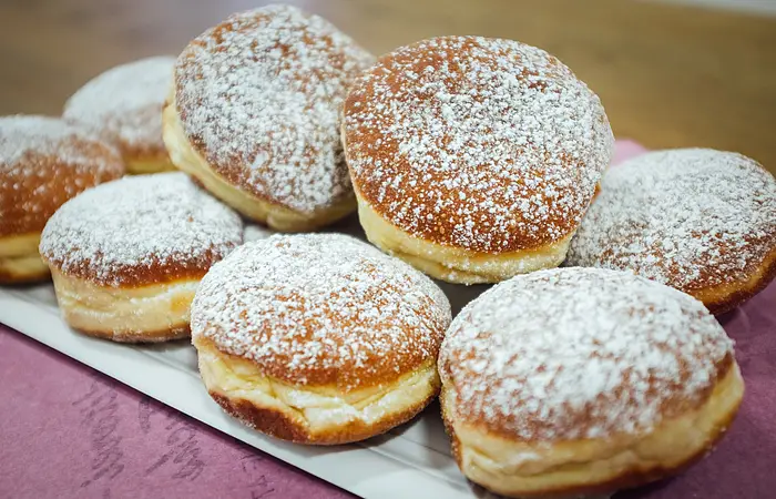 Auf einem weißen Teller sind zehn Krapfen mit Hagebuttenfüllung und Puderzucker geschlichtet. Der Teller steht auf einem Tisch mit rosa Tischdecke.