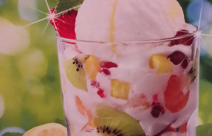 Ein Glasbecher mit verschiedenen Kugeln Eis und leckeren Früchten, wie Kiwi, Erdbeere, Weintraube gefüllt, steht auf einem Tisch. Im Hintergrund ist eine aufgeschnittene Zitrone zu sehen.