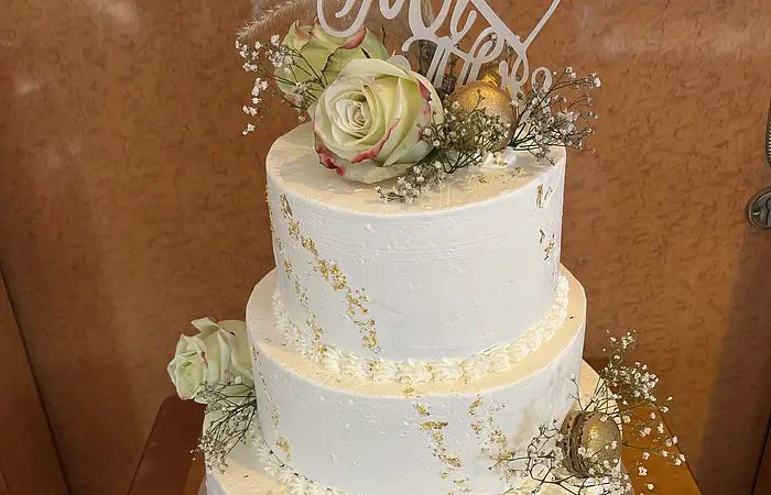 Auf einem Holztisch steht eine dreistöckige Hochzeitstorte, die mit weißer Creme überzogen ist. Die Torte ist elegant mit Blumendekorationen verziert, die ihr ein festliches und ansprechendes Aussehen verleihen.