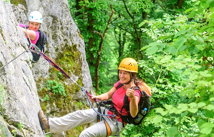 Eine Kletterin mit blonden Haaren hängt gesichert locker am Seil mitten am Felsen. Eine zweite ist schon ein Stück weiter und grinst herüber.