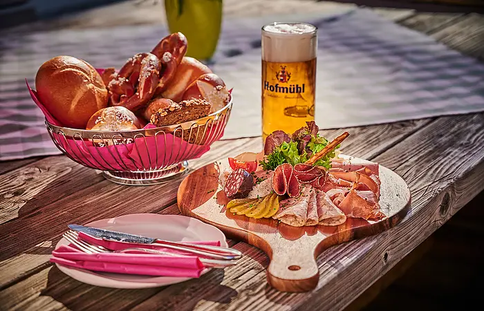 Ein gemütliche Brotzeit im Biergarten.
Hierzu wird ein Brotzeitbrettl mit einem Korb Brezen/Semmeln und einem Bier serviert.