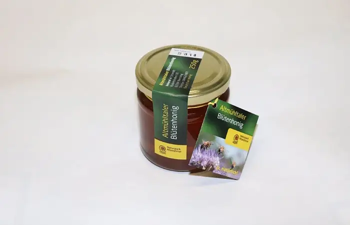 Ein Glas Altmühltaler Blütenhonig mit goldenem Deckel und grünem Etikett auf weißem Hintergrund.