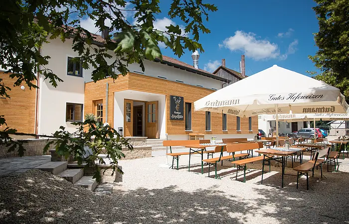 Vor dem Eingang ist ein schön angelegter Biergarten mit Biertischgarnituren und hellen Sonnenschirmen zu sehen. Das Haus ist mit Holzelementen und weißen Wänden versehen.