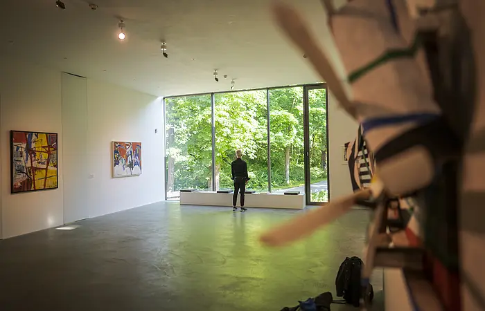 Ein Mann steht vor einer großen hellen Glaswand. Rechts und Links sind Exponate an der Wand zu erkennen.