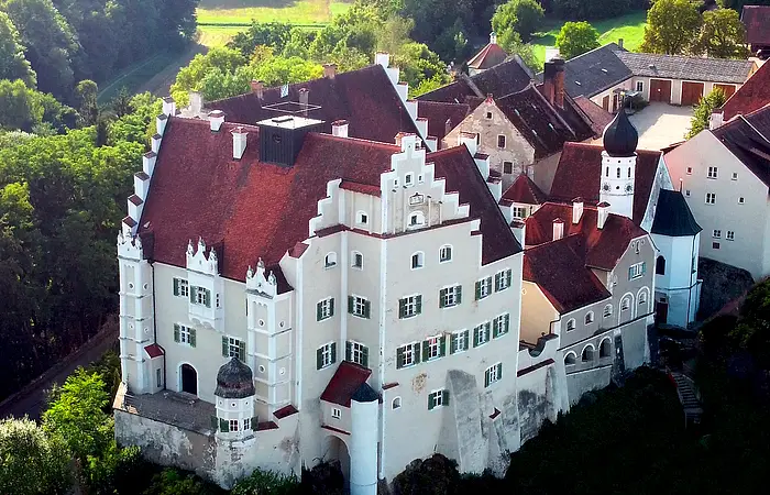 Luftaufnahme eines historischen Schlosses mit roten Dächern, weißen Mauern und umliegendem Wald.