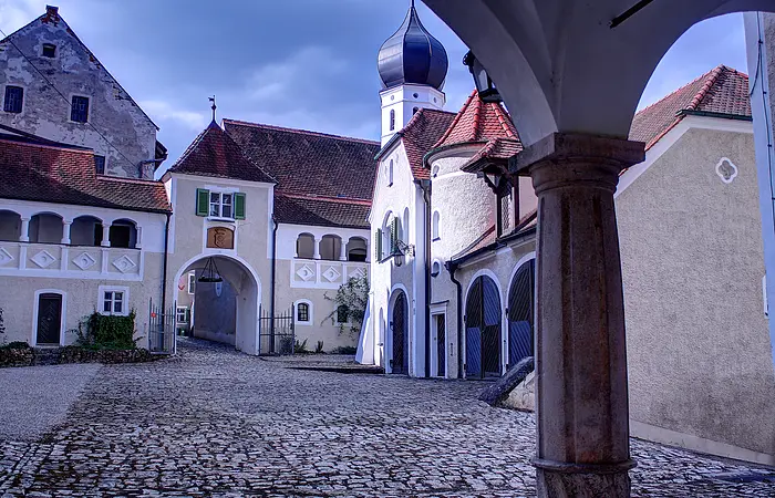 Innenhof eines historischen Gebäudekomplexes mit Kopfsteinpflaster und Torbogen unter bewölktem Himmel.