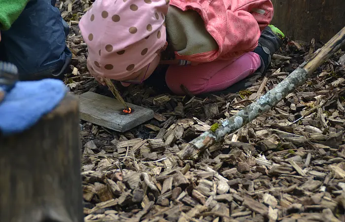 In Kind kniet am Boden und pustet auf ein glimmendes Stück Holz