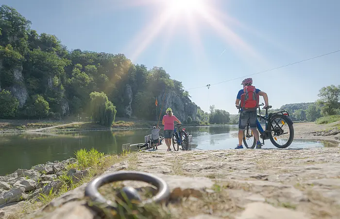 Personen mit Fahrrädern stehen an einem Flussufer mit bewaldeten Felsen im Hintergrund bei Sonnenschein.