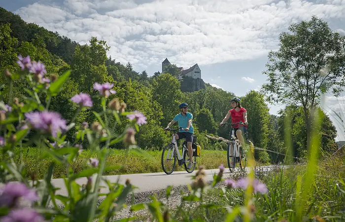 Zwei Radler, eine Frau mit weißem Helm und roten Shirt und ein Mann, neben ihr, ebenfalls mit Helm und blauen Shirt sind auf dem Radweg unterwegs. Die Zwei radeln und hinter ihnen ragt die Burg Prunn, die umgeben von Wäldern auf einem Felsen. Der Himmel scheint blau, nur ein paar Wolken verirren sich.