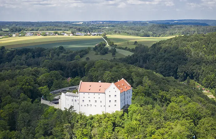 Luftaufnahme eines weißen Schlosses mit rotem Dach, umgeben von Wald und Feldern unter blauem Himmel.
