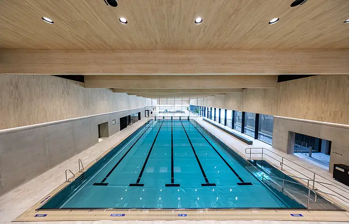 Innenaufnahme eines Schwimmbeckens mit Holzdecke und großen Fenstern an der rechten Seite