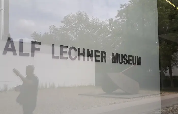 Glasfassade mit Schriftzug "Alf Lechner Museum", Spiegelung von Bäumen und einer Person.