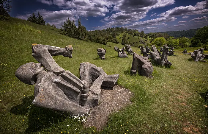Die Fotographie zeigt ein Skulpturenfeld, welches auf einer Wiese in Hanglage gelegen ist. Die abstrahierten, menschenartigen Figuren sind aus Beton gegossen und stellen Kämpfer und Opfer in verschiedenen Positionen dar. Die Gesamtheit des Figurenfeldes erinnert an ein Schlachtfeld. Der Hintergrund zeigt eine karge Berglandschaft.