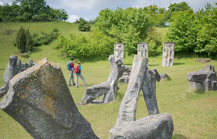 Auf den Wiesen im Hessental sind 78 überlebensgroße Plastiken, die zu einem fesselnden Mahnmal gegen Krieg und Gewalt geschaffen wurden, zu sehen. Inmitten dieses sind zwei Wanderer mit Rucksäcken unterwegs.