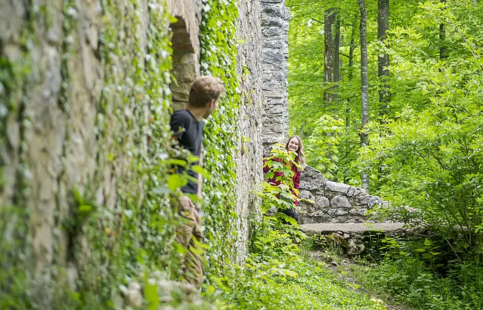 Zwei Personen schauen um eine mit Efeu bewachsene Steinmauer in einem Waldgebiet.