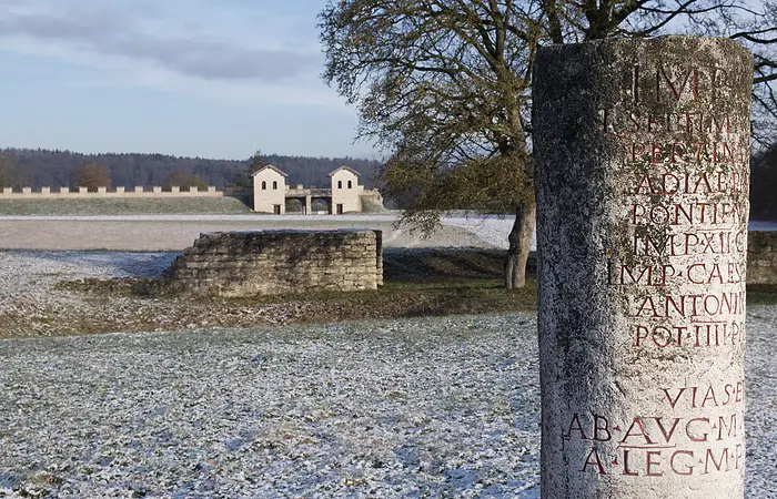 Im Vordergrund befindet sich ein römischer Meilenstein mit Entfernungsangaben zu den nächsten wichtigen Zielen. Im Hintergrund der winterlichen Landschaft ist die rekonstruierte Toranlage des römischen Kastells Vetoniana zu sehen.