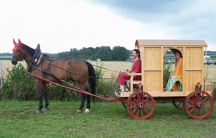 Ein Pferd zieht einen hölzernen Wagen mit zwei Personen in historischer Kleidung über eine Wiese.